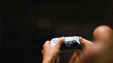 Player holding a white PS5 controller with both thumbs near the analog sticks in front of a TV