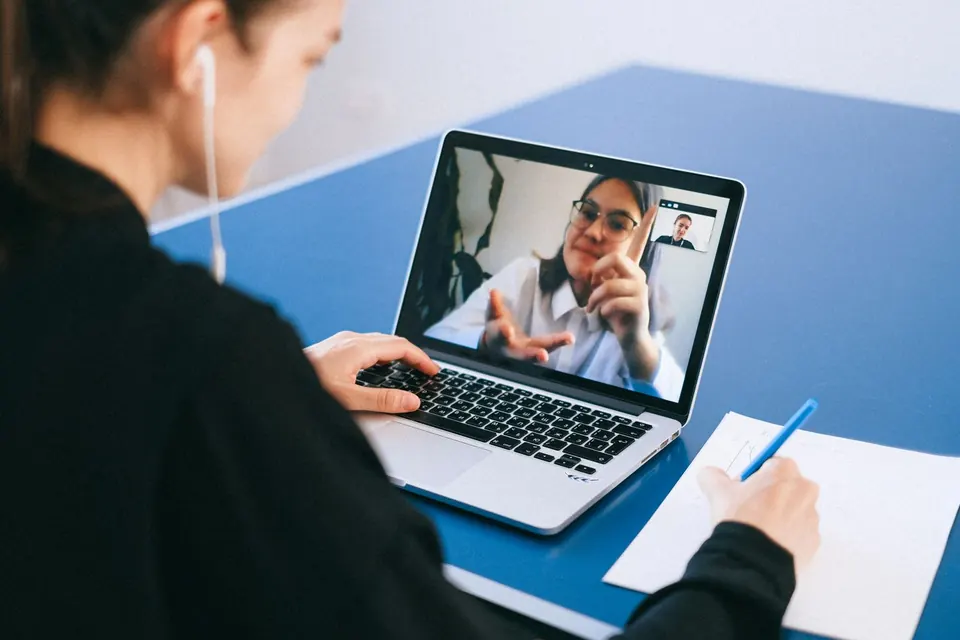 Persona tomando notas en un escritorio durante una videollamada con una colega en la pantalla del portátil