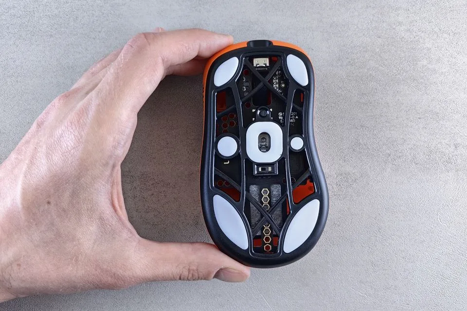 Tangan memegang mouse gaming terbalik memperlihatkan bukaan sensor dan kaki PTFE di bagian bawahnya