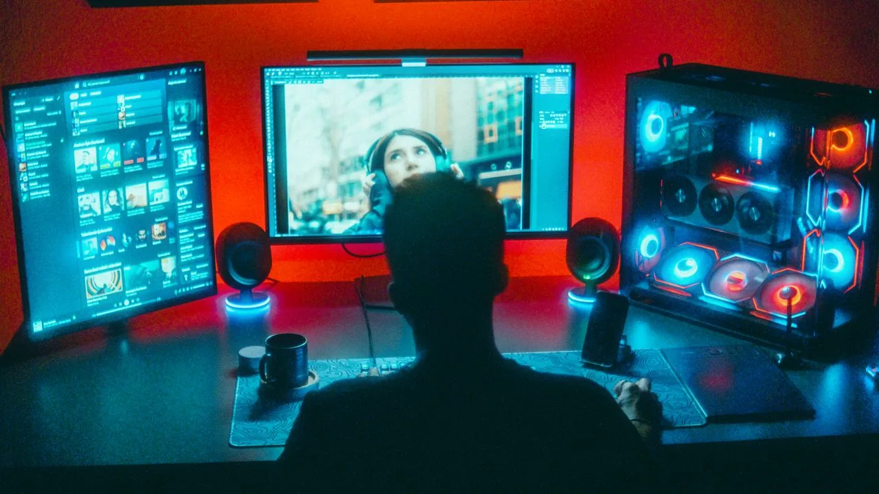 Gamer di meja gelap di depan monitor RGB terang, pola HUD statis kecerahan tinggi yang paling menaikkan risiko burn-in OLED