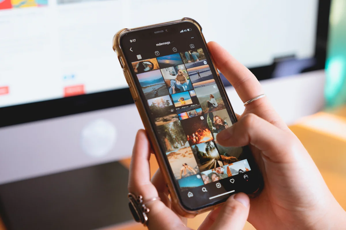 Hands browsing an Instagram photo grid on a phone beside a desktop computer