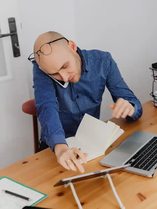 Illustrative stock photo of a man taking a work call beside a laptop and notebook
