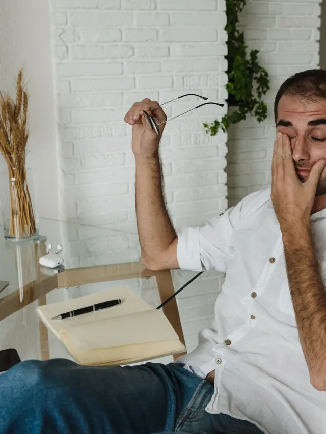 Tired man at a home office desk rubbing his eyes and holding glasses