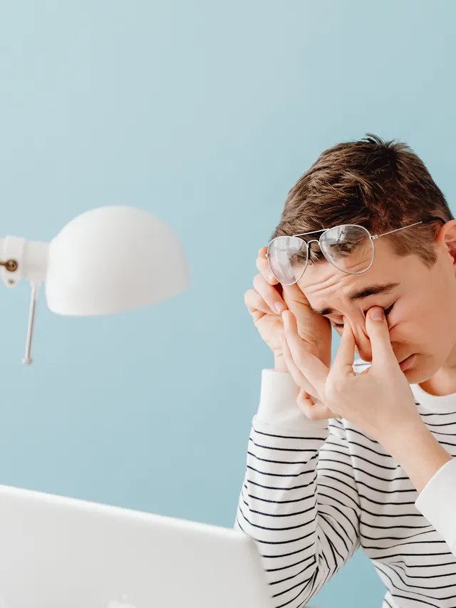 Young man rubbing his eyes at a laptop under a desk lamp