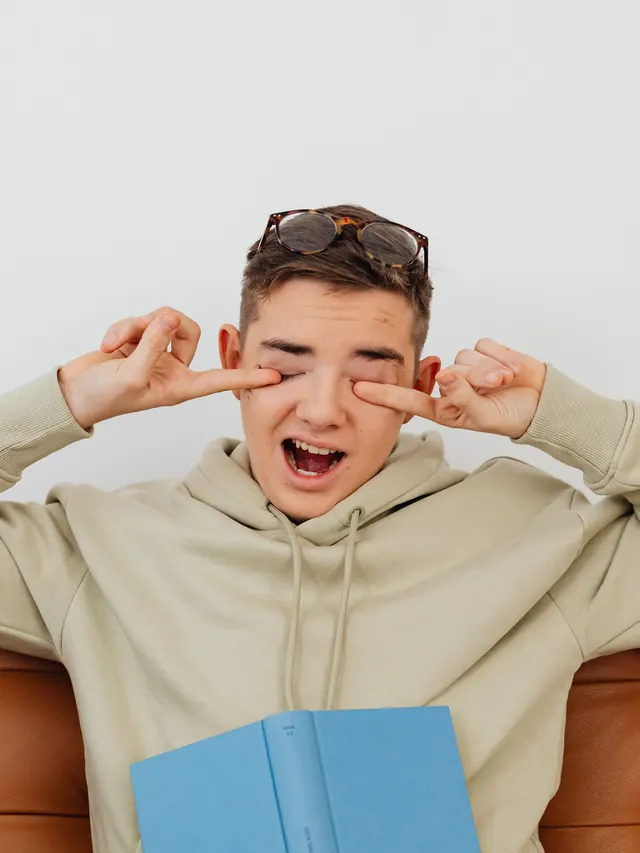 Tired teenager in a hoodie rubbing their eyes on a sofa