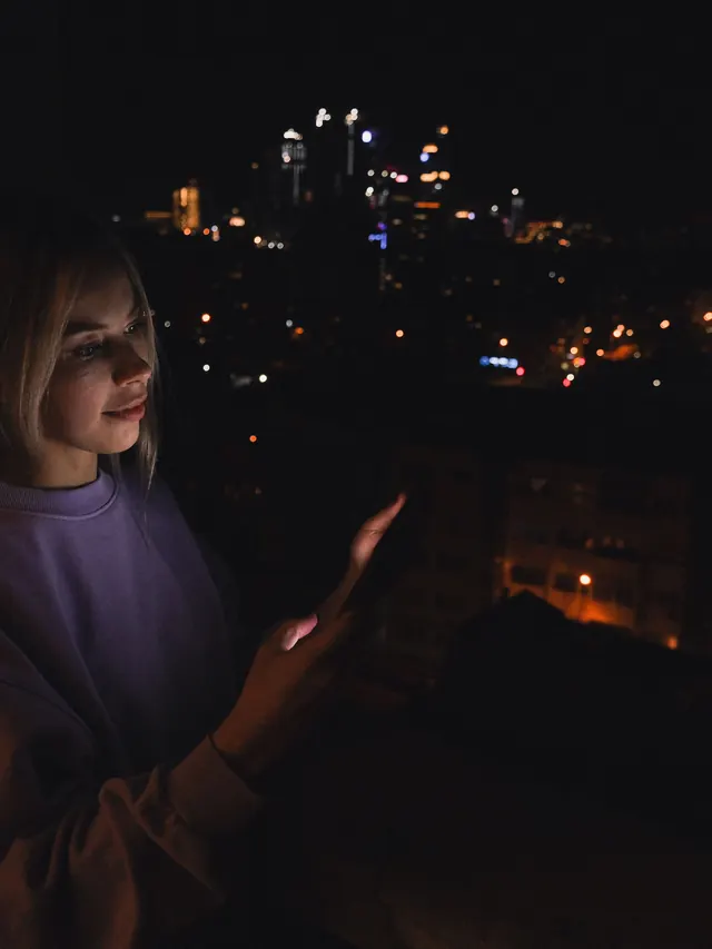 Woman on a balcony at night using a smartphone above city lights