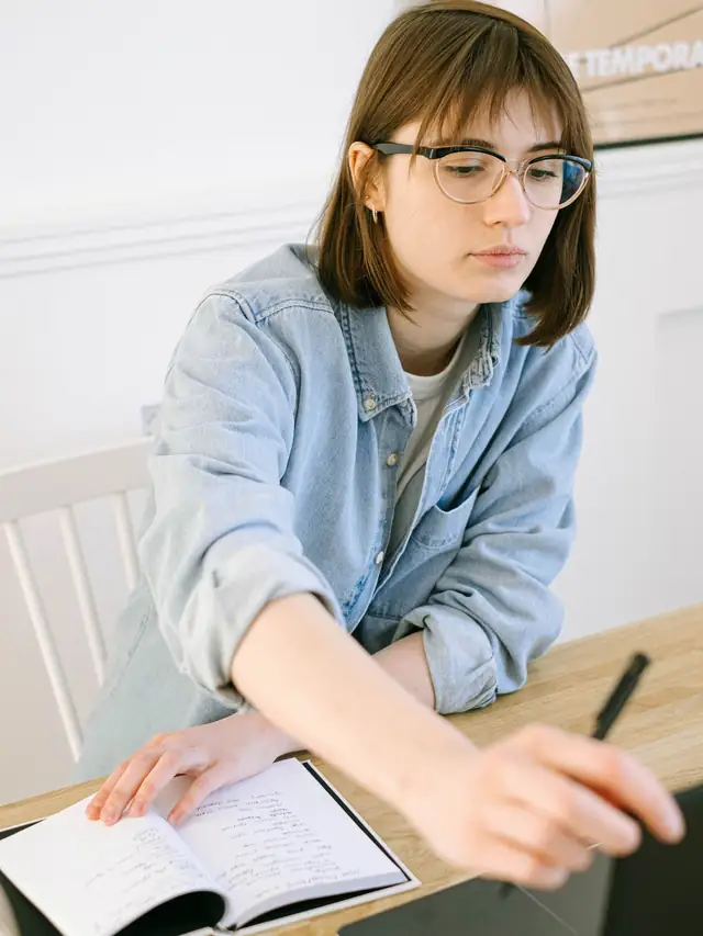 Woman with glasses reviewing notes beside a laptop