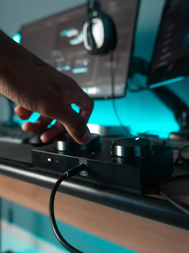 Hand reaching for a control dial at a desk setup with monitors and a headset