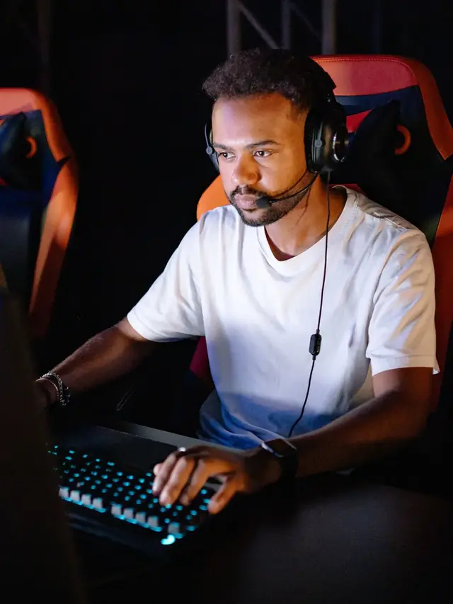Bearded player in a white T-shirt focused on his monitor while running checks at a backlit keyboard