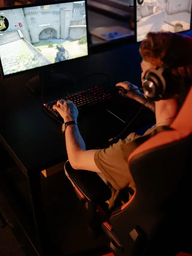 Over-the-shoulder view of a player driving a shooter match on a desktop monitor from a full PC rig