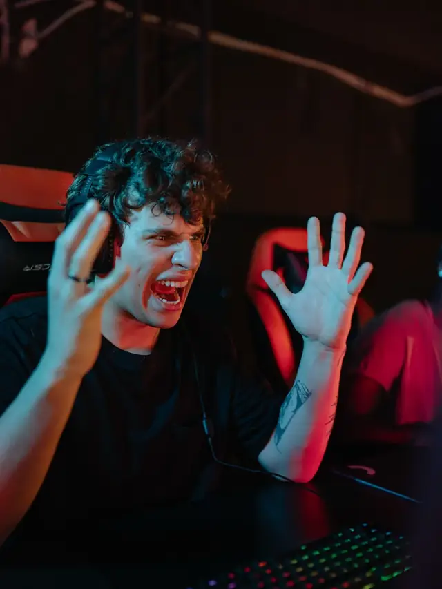 Frustrated curly-haired gamer throwing both hands up at his monitor in red arena light