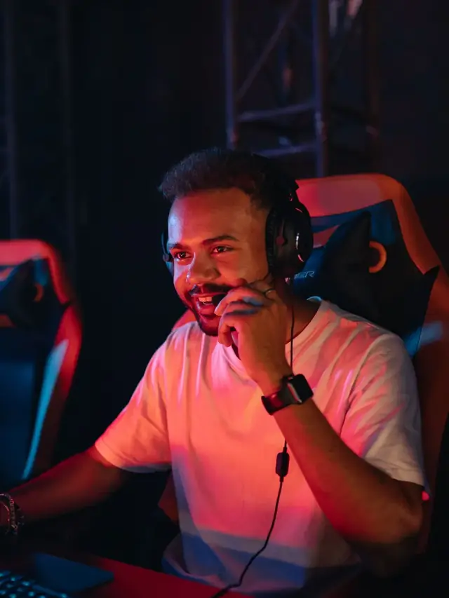 Bearded player in a white T-shirt holding his headset mic and weighing a decision in red light
