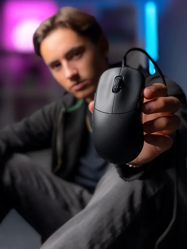Young man holding up the black CHERRY XTRFY M42V2 wired gaming mouse toward the camera in a neon-lit room
