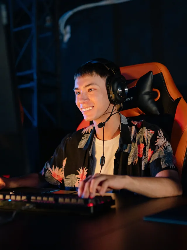 Smiling gamer in a floral shirt and headset with his hands on a keyboard and mouse