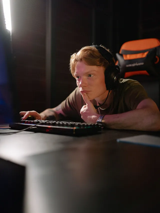 Red-haired gamer in a headset weighing a decision with his hand on his chin at a gaming desk
