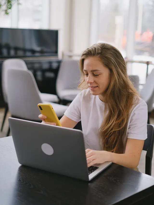Woman researching on a laptop while checking a yellow smartphone