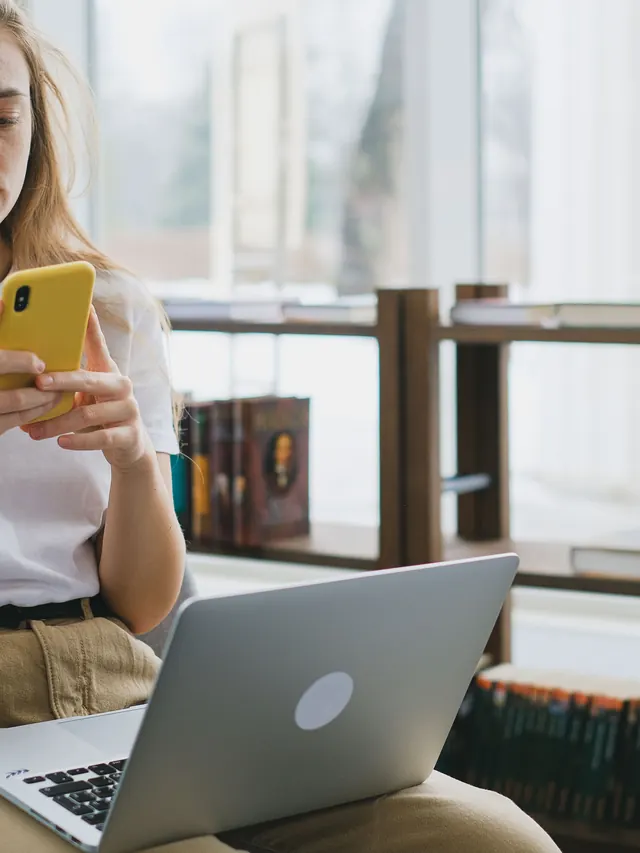 Woman using a yellow smartphone beside a laptop in a bright room