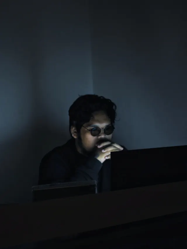Man in eyeglasses weighing information on a computer screen in a dark room