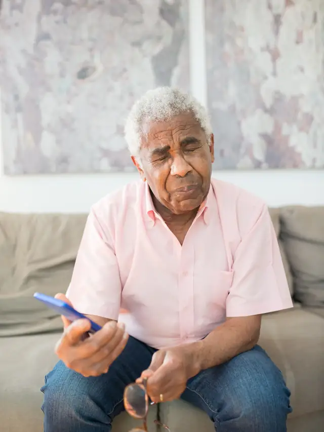 Senior man sitting on a couch holding a smartphone with his eyes closed